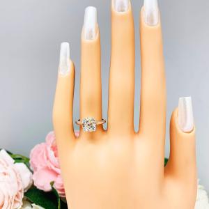 Mannequin hand with long white shimmery nails, displayed against a soft white-gray background with pink and white floral accents. The hand is wearing a sterling silver split-prong cathedral engagement ring set with a near colorless old mine cut cubic zirconia.