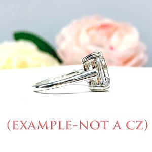 A sterling silver ring with a large, clear, emerald-cut gemstone. The ring is facing toward the right to show the side view and is sitting on a white surface with pink and white flowers in the background. (EXAMPLE - NOT A CZ)