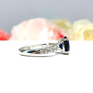 Close-up view of a stunning sterling silver oval-cut blue sapphire engagement ring. The engagement ring features a vibrant dark blue sapphire with pave cubic zirconia side stones.The ring is displayed against a blurred background of soft pink and white roses.