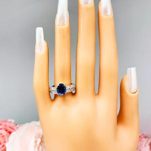 Mannequin hand with long white shimmery nails, displayed against a soft white-gray background with pink and white floral accents. The hand is wearing a stunning sterling silver oval-cut blue sapphire engagement ring and a matching wedding band. The engagement ring features a vibrant dark blue sapphire with pave cubic zirconia side stones. The wedding band is a half-eternity band, also in sterling silver, with a continuous row of smaller white cubic zirconias.
