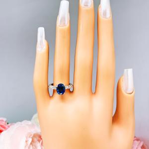 Mannequin hand with long white shimmery nails, displayed against a soft white-gray background with pink and white floral accents. The hand is wearing a stunning sterling silver oval-cut blue sapphire engagement ring. The engagement ring features a vibrant dark blue sapphire with pave cubic zirconia side stones.