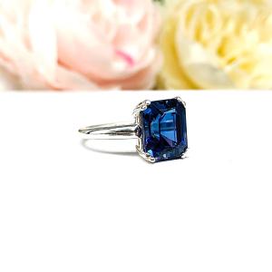 Sterling silver engagement ring featuring a deep blue emerald-cut lab-created blue sapphire in a sleek double-prong setting. The ring is resting on a white surface with soft-focus pink and white flowers.