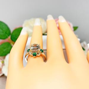 A display hand with long white fingernails laying down and with the top of the hand facing up, displayed against a soft-focus background of pink and white flowers.  The display hand is wearing an Art Deco-style sterling silver ring with a round brilliant near-colorless cubic zirconia center stone, flanked by vivid green lab-created emerald accents.