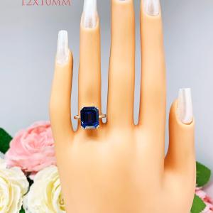 Mannequin hand with long white shimmery nails, displayed against a soft white-gray background with pink and white floral accents. The hand is wearing a sterling silver solitaire ring featuring a deep blue emerald-cut lab-created blue sapphire in a sleek double-prong setting. The text '12X10MM' appears in the image.