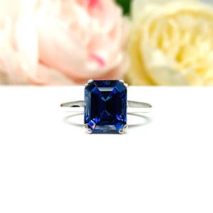 Sterling silver engagement ring featuring a deep blue emerald-cut lab-created blue sapphire in a sleek double-prong setting. The ring is resting on a white surface with soft-focus pink and white flowers.