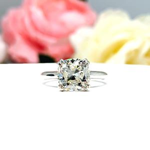 A sterling silver split-prong cathedral engagement ring set with a near colorless old mine cut cubic zirconia. The ring is set against a white background with a blurred image of pink and white flowers in the background.