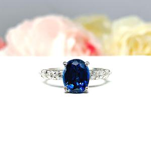 Close-up view of a stunning sterling silver oval-cut blue sapphire engagement ring. The engagement ring features a vibrant dark blue sapphire with pave cubic zirconia side stones.The ring is displayed against a blurred background of soft pink and white roses.