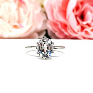Sterling silver ring with an oval lab created white sapphire set in a 6-prong setting set against a white surface with soft-focus pink flowers in the background.