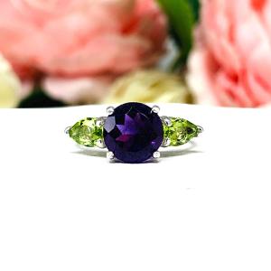 Sterling silver ring featuring a vibrant 8mm round African amethyst flanked by two pear-cut peridot side stones. The rich purple and bright green gemstones create a striking contrast, set against a soft floral background that enhances the ring’s natural elegance.