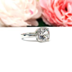 A sterling silver ring with a large, cushion-cut lab white sapphire. The stone is set in a bezel prong setting, and the band is rounded and polished. The background features soft pink and white flowers, creating a romantic aesthetic.