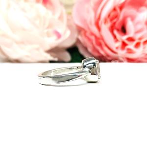 A sterling silver ring with a large, cushion-cut lab white sapphire. The stone is set in a bezel prong setting, and the band is rounded and polished. The background features soft pink and white flowers, creating a romantic aesthetic.