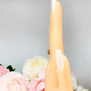 Mannequin hand with long white shimmery nails, displayed against a soft white-gray background with pink and white floral accents. The hand is wearing a sterling silver ring with a large, cushion-cut lab white sapphire. The stone is set in a bezel prong setting, and the band is rounded and polished.