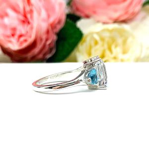 A sterling silver 3-stone ring featuring an 11x9mm lab created white sapphire flanked by two 5mm swiss blue topaz gemstones. Displayed on a white surface with a floral background.