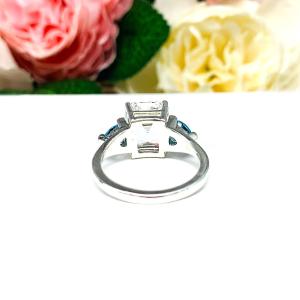 A sterling silver 3-stone ring featuring an 11x9mm lab created white sapphire flanked by two 5mm swiss blue topaz gemstones. Displayed on a white surface with a floral background.