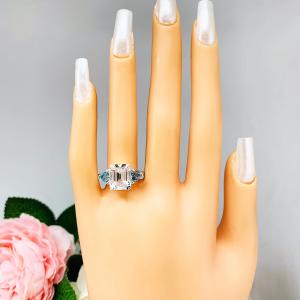 Mannequin hand with long white shimmery nails, displayed against a soft white-gray background with pink and white floral accents. The hand is wearing a sterling silver 3-stone ring featuring an 11x9mm lab created white sapphire flanked by two 5mm swiss blue topaz gemstones.