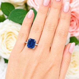 A woman's hand wearing a sterling silver double prong solitaire ring featuring a 12x10mm emerald cut lab blue sapphire. Model image for size and styling reference — not a photo of the actual ring.