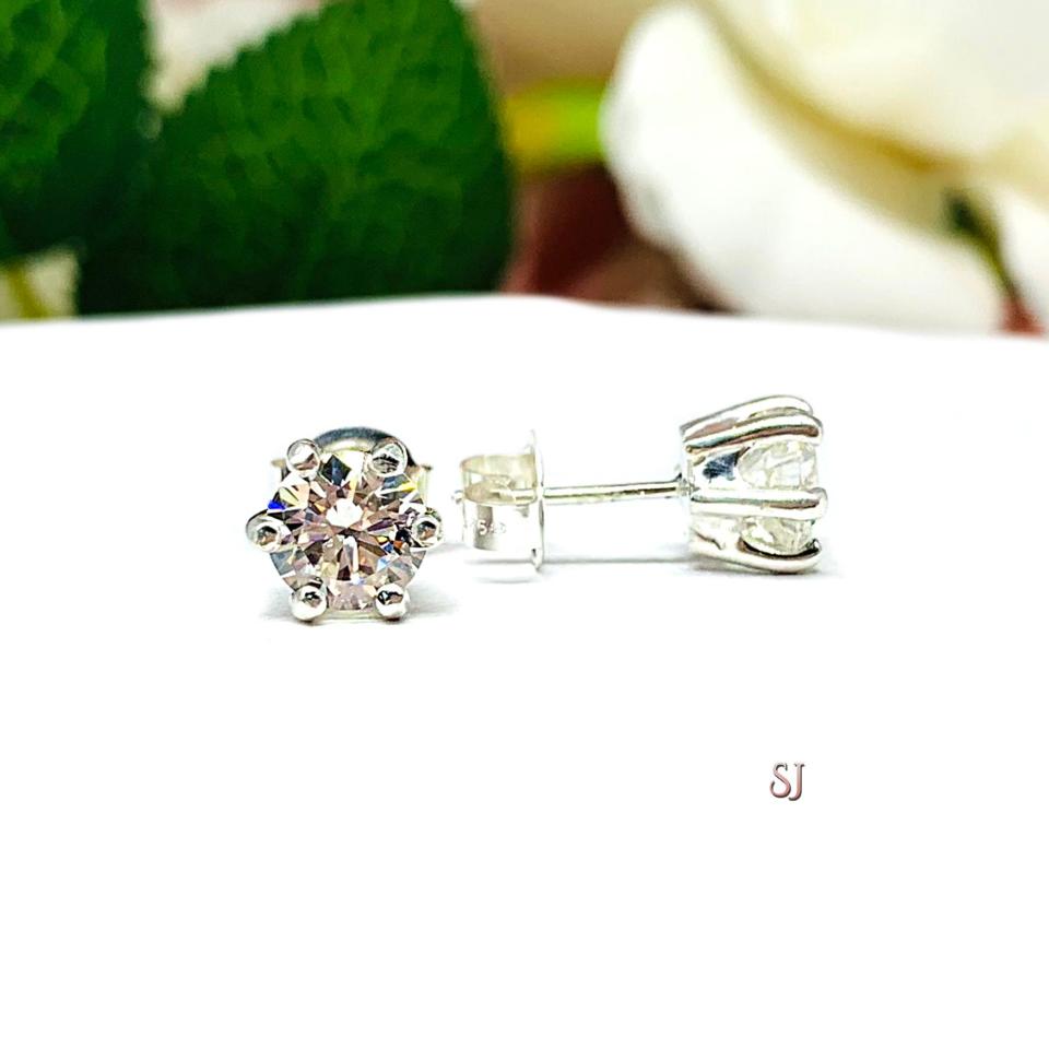 A pair of silver stud earrings set with round brilliant cut 'near colorless or 'G color' cubic zirconias.  The 6 prong earrings are on a white surface with the left earring facing forward and the right earring facing to the right to showcase the brilliant cutting of the czs, center post and butterfly friction clasp. The text 'SJ' appears in pink to the right, below the earrings.