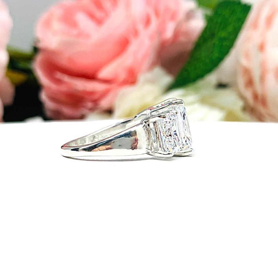 Sterling silver 3-stone engagement ring featuring an 8x6mm-10x8mm radiant cut cubic zirconia center stone with two smaller radiant CZ side stones. The highly polished wide band tapers for comfort. Displayed on a white surface with soft floral background.