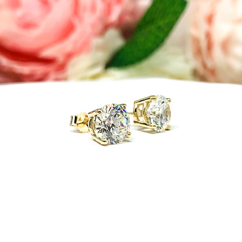 A pair of yellow gold stud earrings set with round brilliant cut 'near colorless or 'G color' cubic zirconias.  The four prong basket earrings are on a white surface with both earrings facing slightly to the right to showcase the brilliant cutting of the czs, center post and butterfly friction clasps.