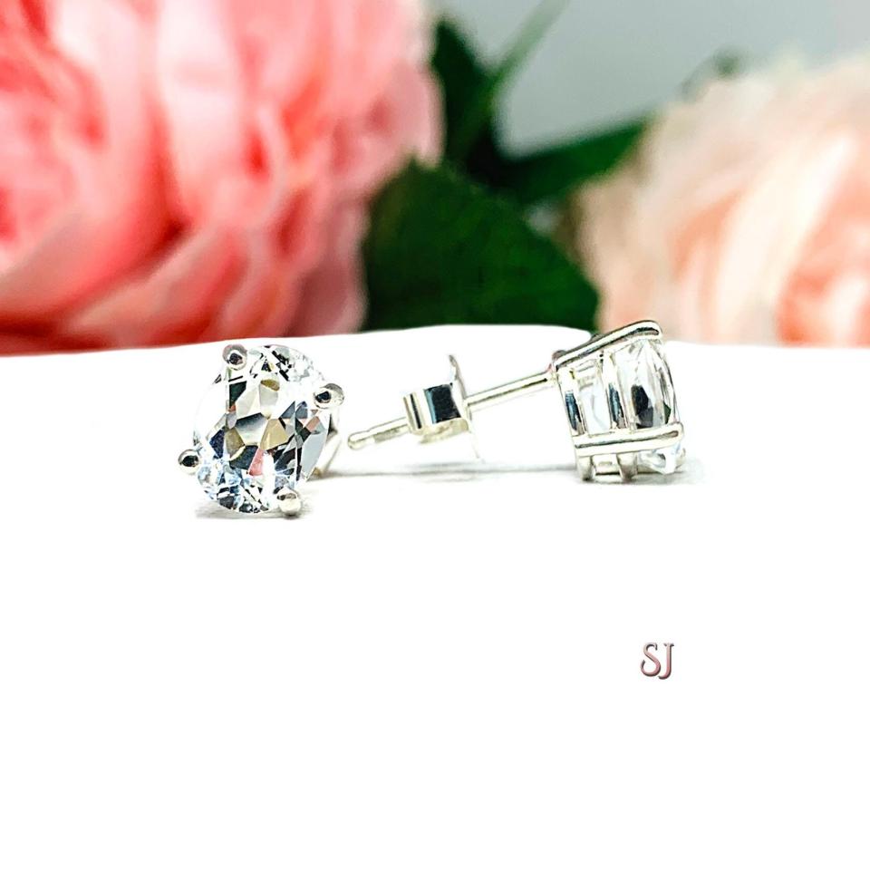 A pair of silver basket earrings set with oval natural white topaz gemstones, displayed with one earring facing the front to showcase clear clarity of the gemstones and one facing to the side to display that the post sits at the top of the earring and the butterfly friction clasp. The earrings are shown on a white background.