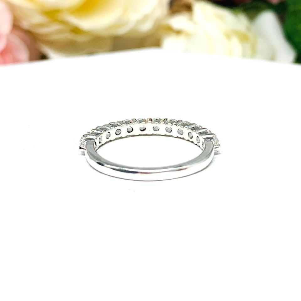 Sterling silver band with 12 round 2.5mm white topaz gemstones set in a shared prong setting. The band is on a white surface with pink and white flowers in the background.
