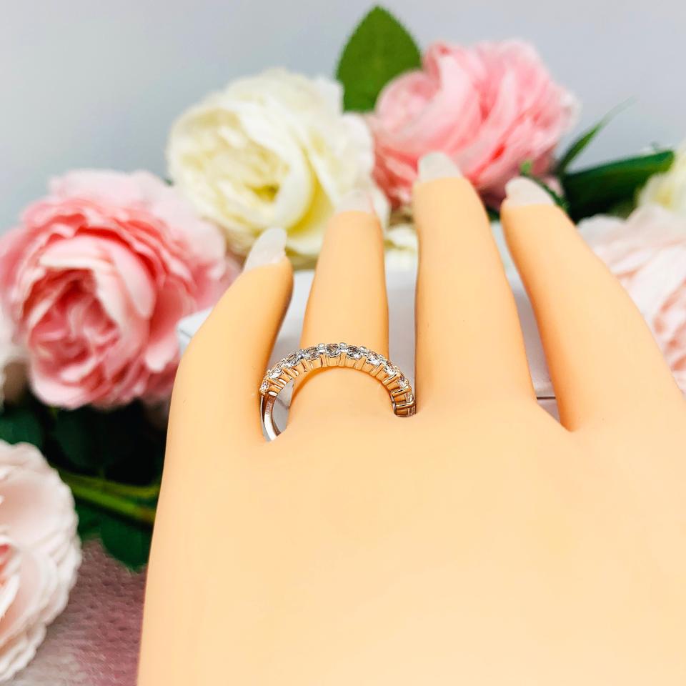 Mannequin hand with long white shimmery nails, displayed against a soft white-gray background with pink and white floral accents. The hand is wearing a sterling silver band with 12 round 2.5mm white topaz gemstones set in a shared prong setting.