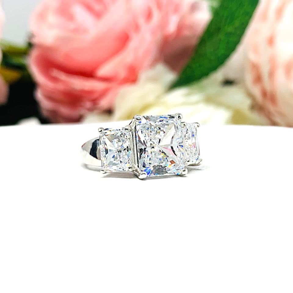 Sterling silver 3-stone engagement ring featuring an 8x6mm-10x8mm radiant cut cubic zirconia center stone with two smaller radiant CZ side stones. The highly polished wide band tapers for comfort. Displayed on a white surface with soft floral background.