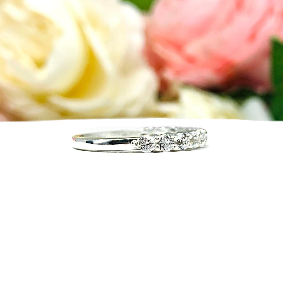 Sterling silver band with 12 round 2.5mm white topaz gemstones set in a shared prong setting. The band is on a white surface with pink and white flowers in the background.