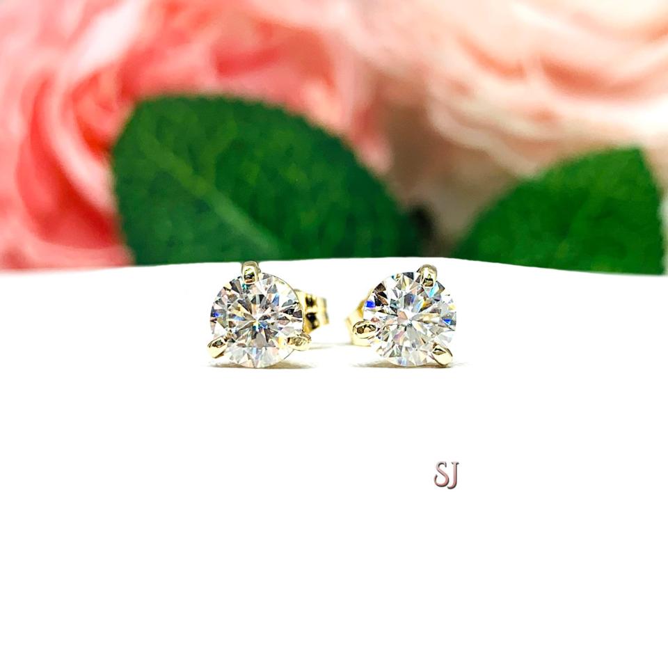 A pair of yellow gold martini stud earrings with round brilliant moissanites. The earrings are on a white background with both earrings facing forward to showcase the sparkling stones. The text 'SJ' is visible in the lower right corner of the image.
