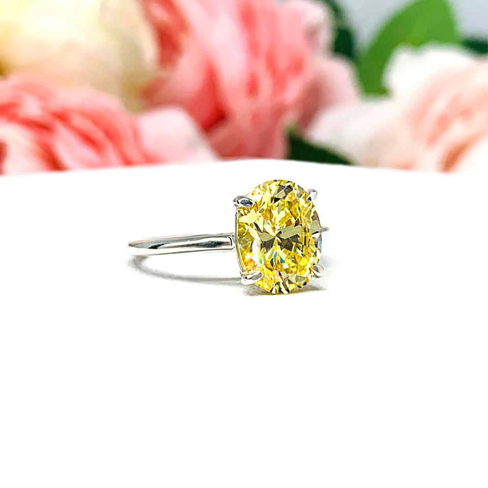A sterling silver claw prong solitaire cathedral engagement ring featuring a well-faceted, canary yellow 10x8mm cz. Shown on a white surface with a pastel floral background.