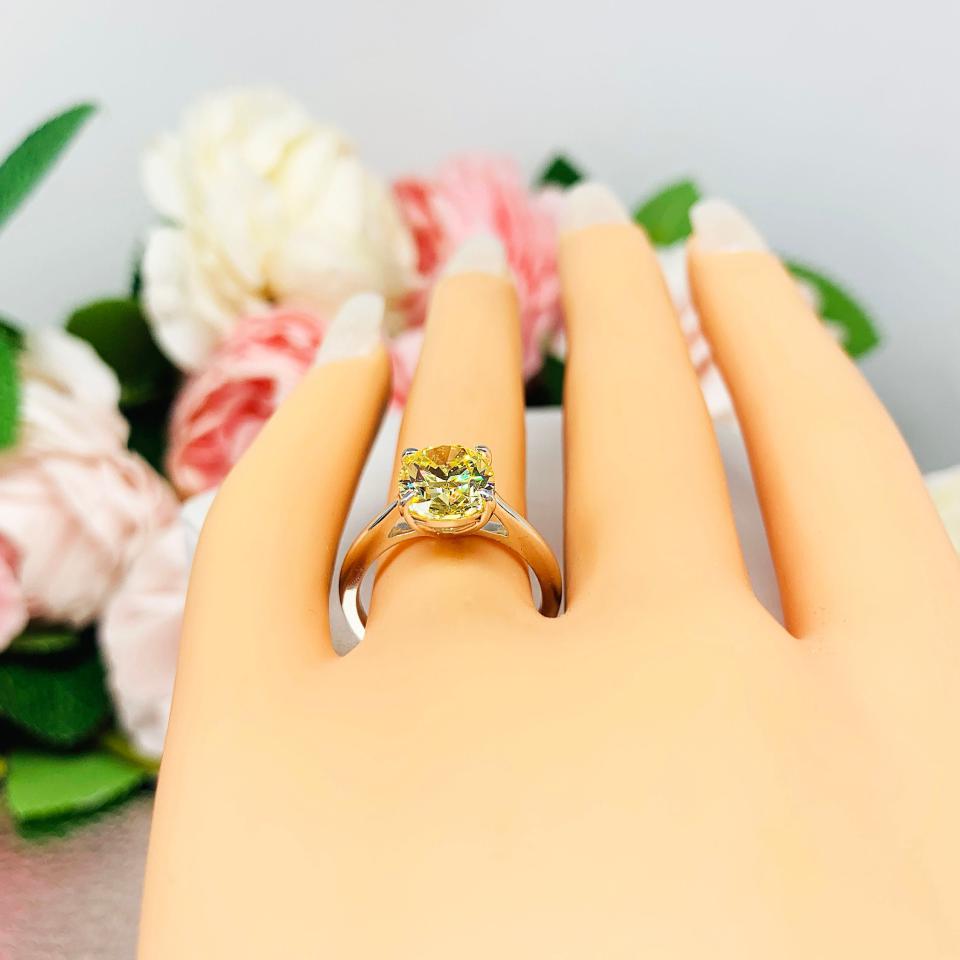 Mannequin hand with long white shimmery nails, displayed against a soft white-gray background with pink and white floral accents. The hand is wearing a sterling silver claw prong solitaire cathedral engagement ring featuring a well-faceted, canary yellow 10x8mm cz.