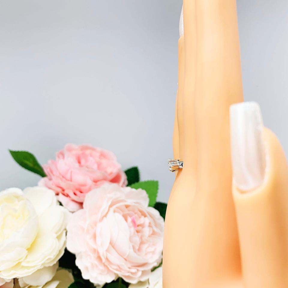 Mannequin hand with long white shimmery nails, displayed against a soft white-gray background with pink and white floral accents. The hand is wearing a 6-prong sterling silver, horizontal east west set engagement ring featuring a precision-faceted near colorless (G/H color) marquise cubic zirconia.