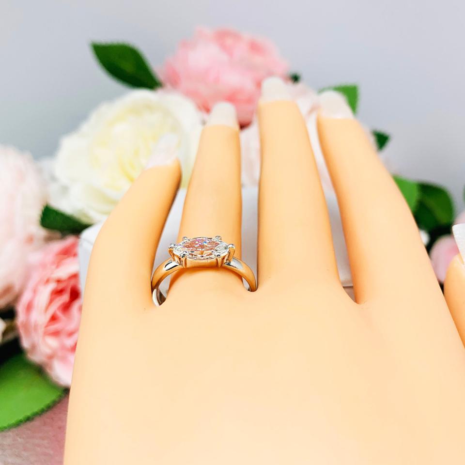 Mannequin hand with long white shimmery nails, displayed against a soft white-gray background with pink and white floral accents. The hand is wearing a 6-prong sterling silver, horizontal east west set engagement ring featuring a precision-faceted near colorless (G/H color) marquise cubic zirconia.