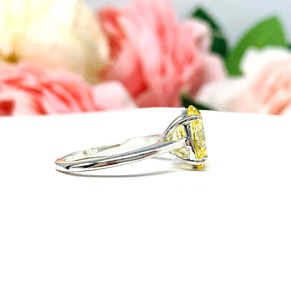 A sterling silver claw prong solitaire cathedral engagement ring featuring a well-faceted, canary yellow 10x8mm cz. Shown on a white surface with a pastel floral background.