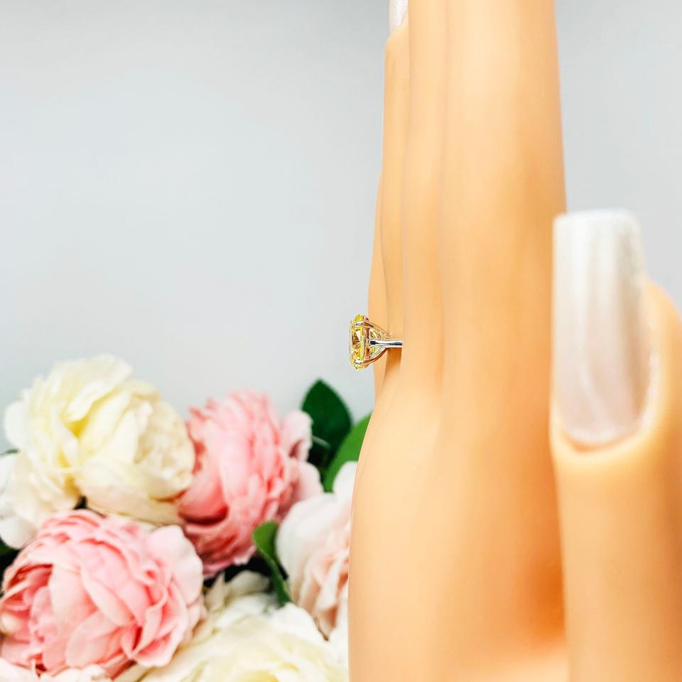 Mannequin hand with long white shimmery nails, displayed against a soft white-gray background with pink and white floral accents. The hand is wearing a sterling silver claw prong solitaire cathedral engagement ring featuring a well-faceted, canary yellow 10x8mm cz.