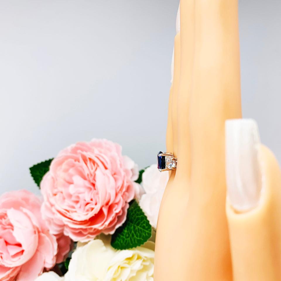 Mannequin hand with long white shimmery nails, displayed against a soft white-gray background with pink and white floral accents. The hand is wearing a sterling silver 3-stone ring with a round lab-created blue sapphire center flanked by smaller round brilliant cut cubic zirconias on each side.