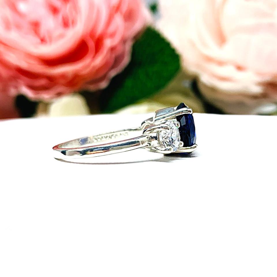 Sterling silver 3-stone ring with a round lab-created blue sapphire center flanked by smaller round brilliant cut cubic zirconias on each side. The ring is set against a white surface with a background of pink and white flowers.