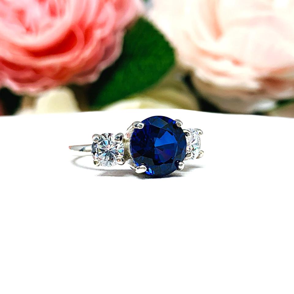Sterling silver 3-stone ring with a round lab-created blue sapphire center flanked by smaller round brilliant cut cubic zirconias on each side. The ring is set against a white surface with a background of pink and white flowers.
