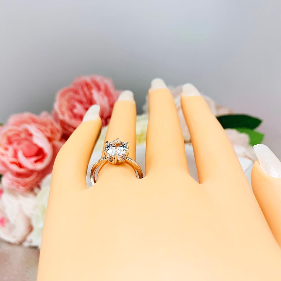 Mannequin hand with long white shimmery nails, displayed against a soft white-gray background with pink and white floral accents. The hand is wearing a sterling silver ring with an oval lab created white sapphire set in a 6-prong setting.