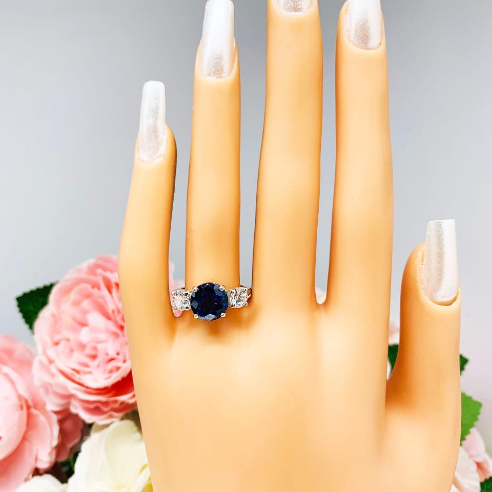 Mannequin hand with long white shimmery nails, displayed against a soft white-gray background with pink and white floral accents. The hand is wearing a sterling silver 3-stone ring with a round lab-created blue sapphire center flanked by smaller round brilliant cut cubic zirconias on each side.