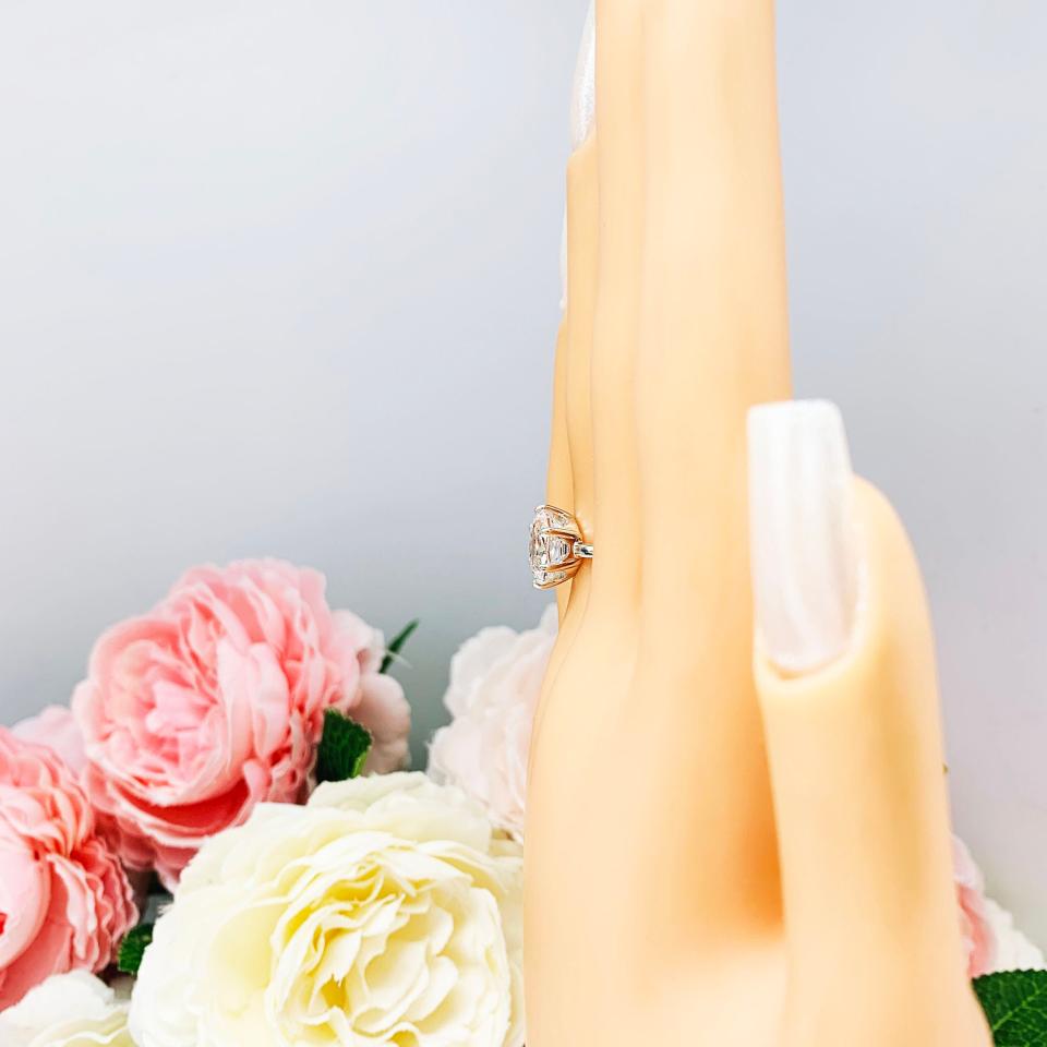 Mannequin hand with long white shimmery nails, displayed against a soft white-gray background with pink and white floral accents. The hand is wearing a sterling silver ring with an oval lab created white sapphire set in a 6-prong setting.