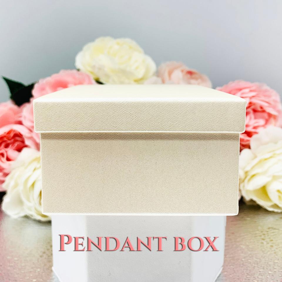 An ivory color square, cardboard box with a textured surface. The box is sitting on a white surface with the words 'PENDANT BOX' printed in pink letters.