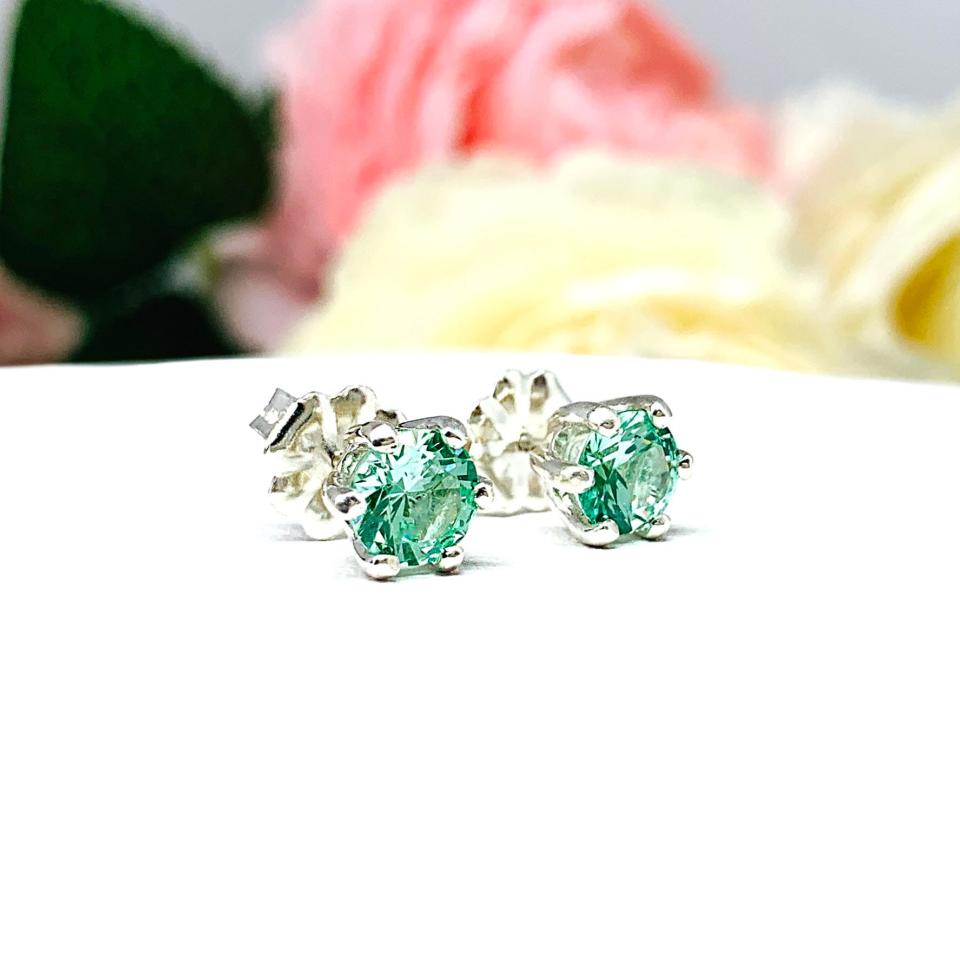 A pair of silver 6 prong earrings set with 6mm round sea foam green lab spinels, displayed with both earrings facing slightly to the right to showcase the centrally located posts and butterfly friction clasps. The earrings are shown on a white background.