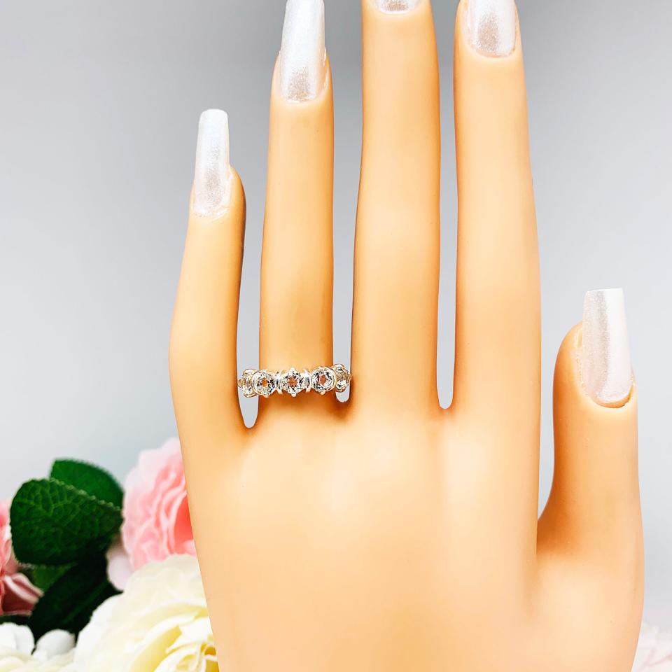 Mannequin hand with long white shimmery nails, displayed against a soft white-gray background with pink and white floral accents. The hand is wearing a sterling silver ring featuring five 4mm round white topaz gemstones in a row. The ring has rounded bars separating each stone.