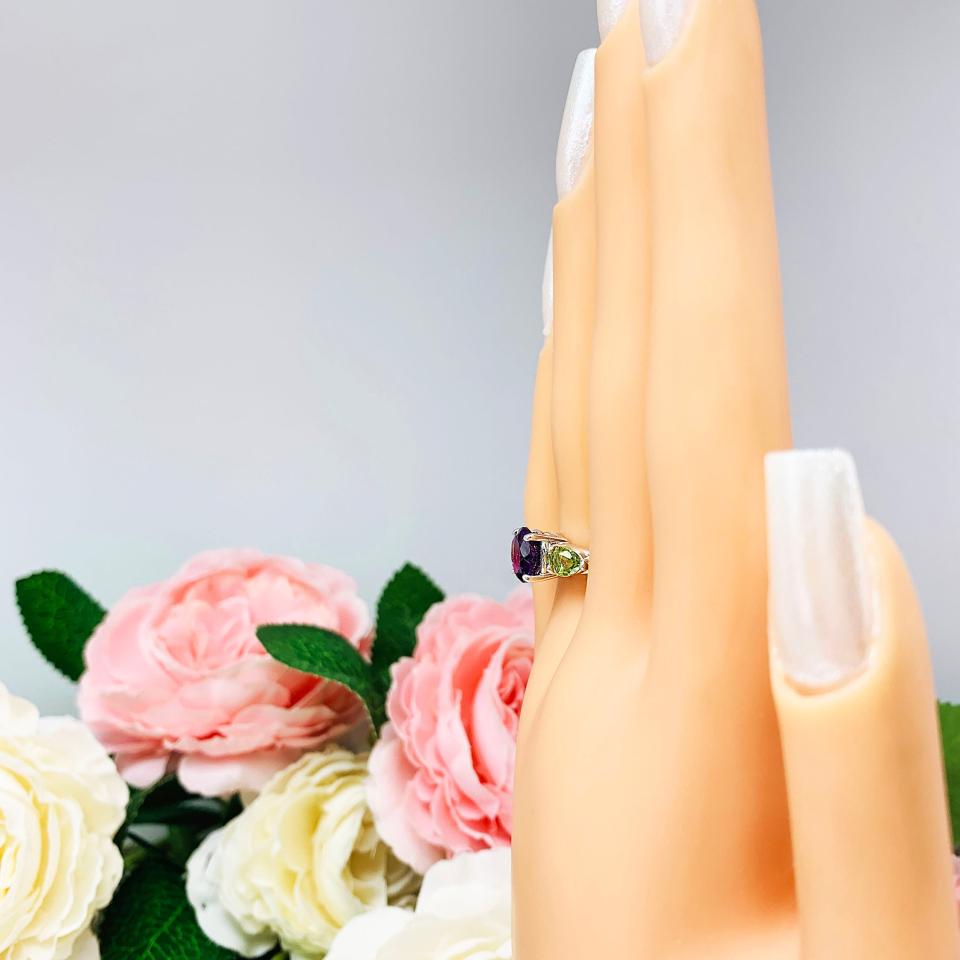 Mannequin hand with long white shimmery nails, displayed against a soft white-gray background with pink and white floral accents. The hand is wearing a sterling silver ring featuring a vibrant 8mm round African amethyst flanked by two pear-cut peridot side stones. The rich purple and bright green gemstones create a striking contrast, set against a soft floral background that enhances the ring’s natural elegance.