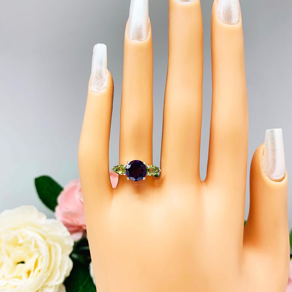 Mannequin hand with long white shimmery nails, displayed against a soft white-gray background with pink and white floral accents. The hand is wearing a sterling silver ring featuring a vibrant 8mm round African amethyst flanked by two pear-cut peridot side stones. The rich purple and bright green gemstones create a striking contrast, set against a soft floral background that enhances the ring’s natural elegance.