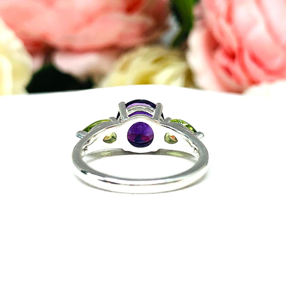 Sterling silver ring featuring a vibrant 8mm round African amethyst flanked by two pear-cut peridot side stones. The rich purple and bright green gemstones create a striking contrast, set against a soft floral background that enhances the ring’s natural elegance.