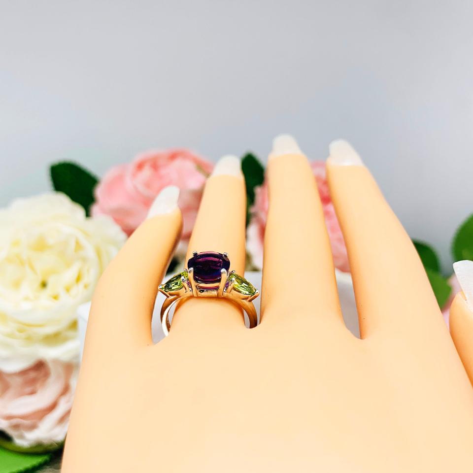 Mannequin hand with long white shimmery nails, displayed against a soft white-gray background with pink and white floral accents. The hand is wearing a sterling silver ring featuring a vibrant 8mm round African amethyst flanked by two pear-cut peridot side stones. The rich purple and bright green gemstones create a striking contrast, set against a soft floral background that enhances the ring’s natural elegance.
