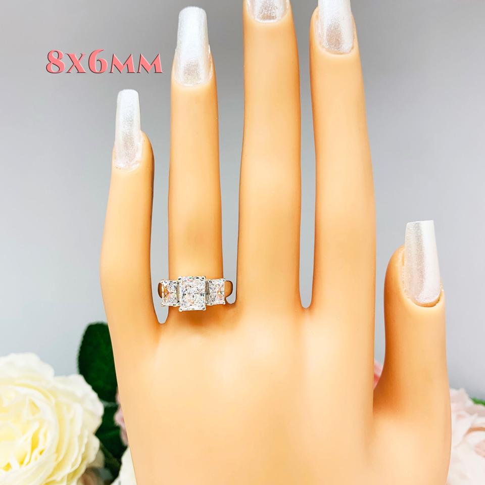 Mannequin hand with long white shimmery nails, displayed against a soft white-gray background with pink and white floral accents. The hand is wearing a sterling silver 3-stone engagement ring featuring an 8x6mm-10x8mm radiant cut cubic zirconia center stone with two smaller radiant CZ side stones. The highly polished wide band tapers for comfort. This variation of the ring features an 8x6mm center stone.