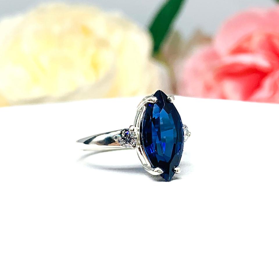 Sterling silver ring showcasing a striking marquise-cut lab-created blue sapphire, it is flanked by two brilliant round cubic zirconia stones. The ring is set against a soft floral backdrop that enhances the ring’s romantic allure.